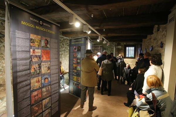 Pubblico alla mostra Mogol: musica e poesia in una sala con soffitto in legno
